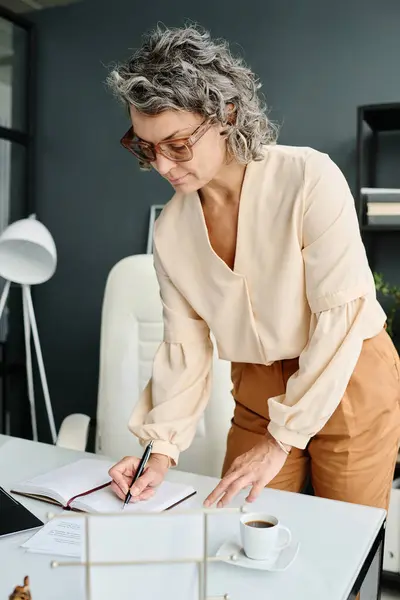 Mature female boss in quiet luxury apparel bending over workplace with open notebook and making working notes in the morning