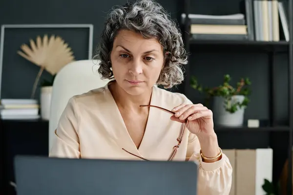 Serious mature female broker or analyst with eyeglasses in hand looking at laptop screen while sitting by workplace and analyzing financial data