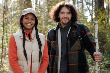 Huş ağacından sopayla gülen genç erkek yürüyüşçü ve hareketli giyimli kız arkadaşı sonbahar ormanında kameranın önünde dikiliyorlar.