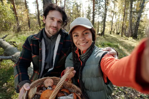 若い異文化カップルのキノコピッカーは 秋の森でカメラを見ながら 携帯電話で写真を撮る — ストック写真