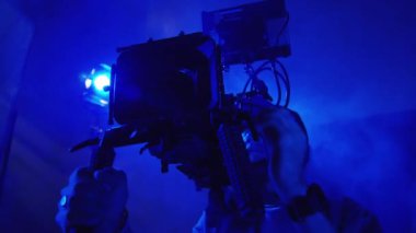 Genç bir erkek kameramanın, profesyonel bir video kamerada, omuz teçhizatında ve karanlık bir stüdyoda, mavi neon ışığı ve dumanla çektiği orta ölçekli bir görüntü.