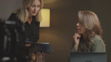 Genç beyaz kadın asistanın, yapımcıyla dizüstü bilgisayarla konuşurken, özet ya da hikaye tahtası gösterirken ve kameraman ekipmanlarını kurarken, stüdyoda reklam filmi çekmeye hazırlanırken orta ölçekli görüntüsü.