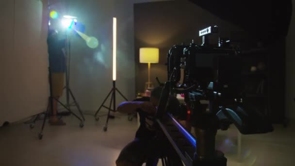 两名男子在黑暗工作室为商业拍摄准备设备 照明技师在三脚架上安装蓝灯和摄象机调整摄像机幻灯片 — 图库视频影像