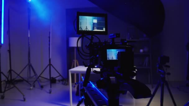 专业摄像机的特写镜头 附加屏幕 在空旷的黑暗制作演播室的玩具平台上拍摄前 背景为蓝色霓虹灯下的桌子和三脚架 — 图库视频影像