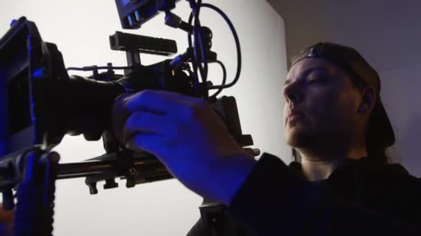 ベースボールキャップのカメラマンのミディアムショットは ショルダーリグにプロのビデオカメラを保持し フレームを調整し 生産クルーにチャットしながら 商業撮影のバックステージで働いています — ストック動画