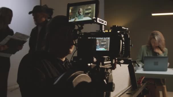 剧组准备在演播室拍摄的中等后台镜头 摄影师在三脚架上安装摄像机 制片人与助手交谈 女主持人与笔记本电脑一起工作 — 图库视频影像