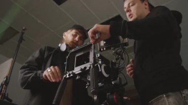 Kafkas erkek görüntü yönetmeni ve teknisyenin stüdyoda yan yana durduğu orta açılı bir çekim, video kameradaki ayarları ayarlamak, objeye bakmak, çekim tekniklerini tartışmak,