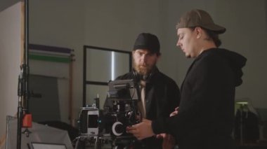 Beyaz tenli erkek kameraman ve asistanın stüdyoda kamerayı çekim için kurarken, önündeki nesnelere bakarken ve operasyonla ilgili konuları tartışırken orta boy görüntüsü.