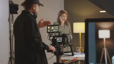 Medya prodüksiyon ekibinin stüdyoda çekim yapmaya hazırlandığı orta ölçekli bir çekim. Tripodlu bir kameraman, modelle çekim açısını tartışıyor ve yapımcı cep telefonuyla konuşuyor.