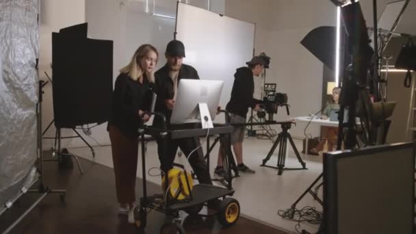 スタジオでのメディア制作クルー撮影のフルショット モデルに向かって人形スライダー上のビデオグラファーの移動カメラ ディレクターと技術者は コンピュータモニター上の映像を検討し 議論しています — ストック動画