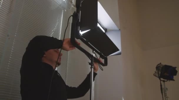野球帽と黒いスウェットシャツに明るいランプをインストールし スタジオで商業撮影する前にビームを調整する白人男性ライトエンジニアのミディアム低角ショット — ストック動画