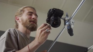 Orta açılı, neşeli, kızıl saçlı, beyaz erkek fotoğrafçı yatay rafa monte edilmiş kameranın düğmesine basarak düz açılı çekim yapar, gülümser ve iş arkadaşıyla sohbet eder.