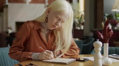 Kafedeki masada oturan albinizmli genç beyaz kadının orta boy portresi öğle yemeğinin bitmesini beklerken çizim yapıyor.