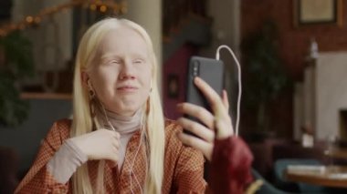 Modern bir restoranda oturmuş video konuşmalarında konuşan, kablo kulaklık takan albino bir kızın orta ölçekli odaklı fotoğrafı.