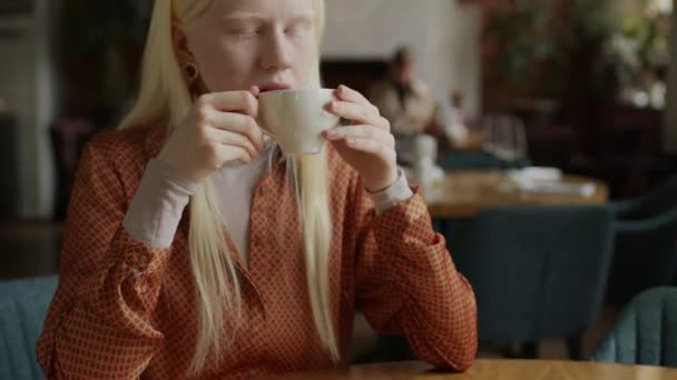 现代咖啡馆里穿着时髦服装的白化病年轻女性坐在餐桌旁喝卡布奇诺的选择性聚焦中特写 — 图库视频影像