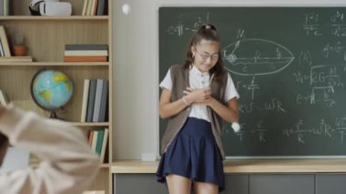 Yavaş çekim orta boy inek gözlüklü bir kızın sınıfta karatahtaya yaslanarak zorbalığa uğradığı sınıf arkadaşlarının ona kağıt toplar fırlattığı fotoğraf.