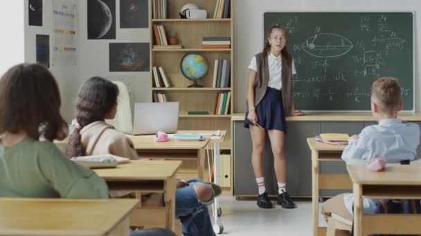 チョークボードに立っているオタクの少女は 彼女のクラスメートが論文を投げ始め 彼女に指を指差し始めたとき 式について話します — ストック動画