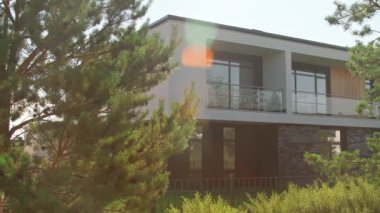 Önünde çam ağaçları büyüyen büyük pencereli ve balkonlu modern evin çekimini yapıyor.
