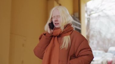 Yavaş çekim. Uzun saçlı genç albino kadın kış günü dışarıda telefonla konuşuyor.