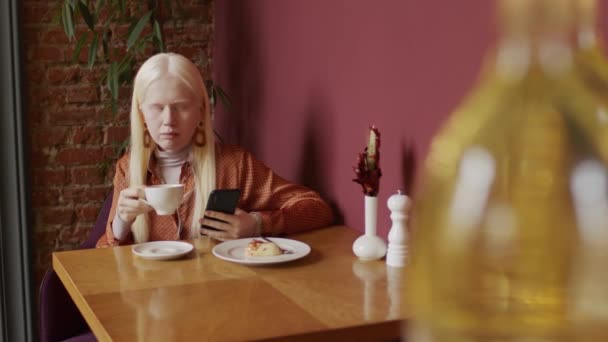 年轻白化病妇女坐在现代咖啡馆餐桌旁 一边喝卡布奇诺 一边用智能手机在网上滚动新闻的选择性聚焦镜头 — 图库视频影像