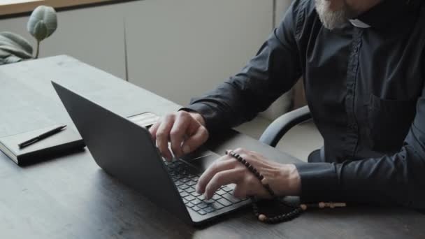 高角度的照片 成熟的白人牧师戴着眼镜和无线耳机 手持念珠 坐在办公室的桌子旁 一边在笔记本电脑上打字 一边打电话 — 图库视频影像