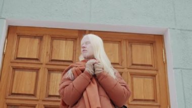 Düşük açılı yavaş çekim. Z albino kızı turuncu kabarık ceketli ve eşarplı ahşap kapılı binanın önünde dikilirken.