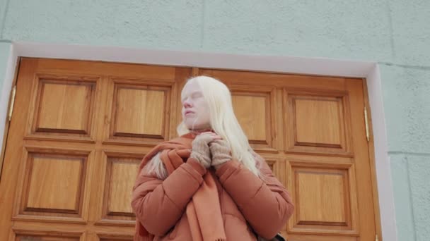 木製のドアとの建物に対して屋外に立っているスカーフが付いているオレンジ色のパッファージのジャケットを身に着けているZアルビノの低い角度のゆっくり動きのショット — ストック動画