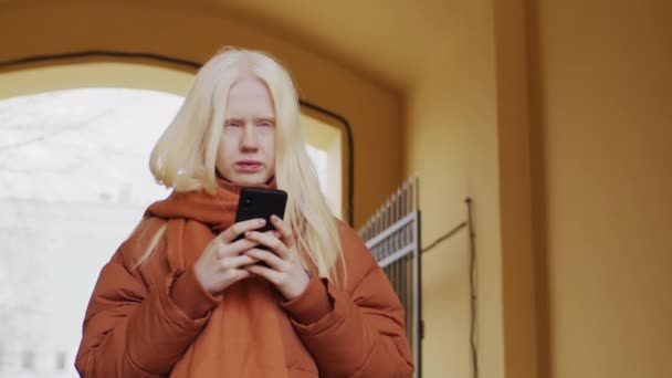 年轻白化病妇女穿着时尚保暖衣服站在户外用智能手机查看某样东西的低角度慢动作肖像 — 图库视频影像