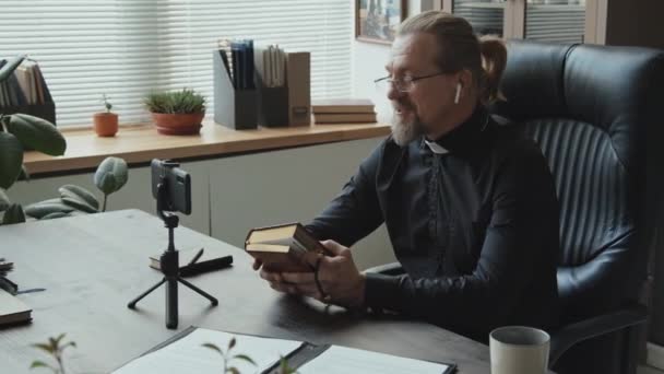 Современный Католический Священник Наушниках Сидит Столом Офисе Записывает Видео Смартфон — стоковое видео
