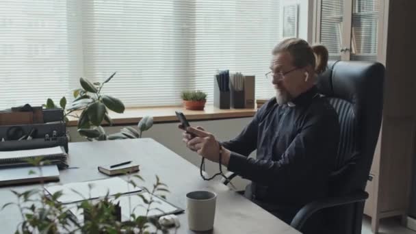 成熟した白人カトリック司祭は ビデオ通話で話している彼のオフィスに座ってワイヤレスイヤホンを身に着けています — ストック動画