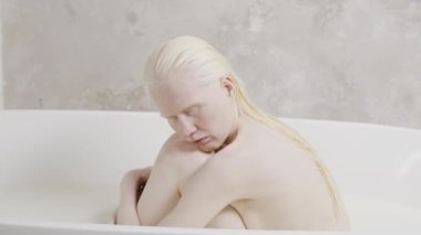 Melankolik albino kadın portresi. Islak saçlı, gözleri kapalı küvette oturmuş, dizlerine sarılıyor.