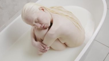 Süt dolu küvette gözleri kapalı, dizlerine sarılan çıplak albino kadının yüksek açılı portresi.