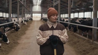 İnek ahırında dikilip kameraya bakan yetişkin, beyaz kadın portresi.