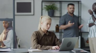 Beyaz kadın yöneticinin dizüstü bilgisayarla çalışırken seçmeli odak noktası. Sinir bozucu erkek iş arkadaşlarının ona iftira atmasını görmezden gelmeye çalışıyor.