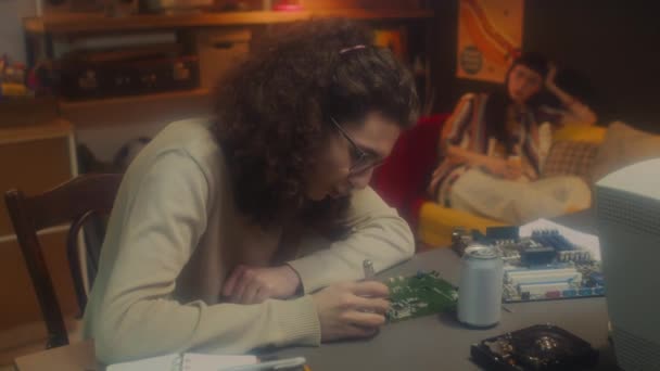 90年代美容美发青年男子坐在办公桌前修理电脑硬件细节 与年轻女子在沙发上放松交谈时的选择性聚焦镜头 — 图库视频影像