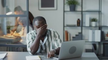 Seçici odak noktası, genç siyahi adamın ofiste otururken dizüstü bilgisayarla uğraşırken yorulması.