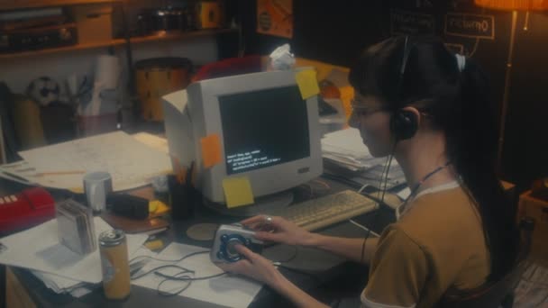 ガレージオフィスでコンピュータで作業するヘッドフォンを身に着けている若い女性プログラマーは 音楽を楽しむために携帯オーディオプレーヤーにカセットを入れて 90年代の美学 — ストック動画