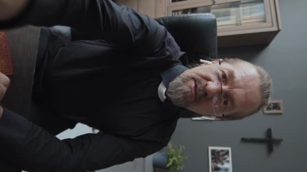 ビデオ通話でスマートフォンを保持しているオフィスに座っているイヤホンを身に着けている成熟したカトリック司祭の垂直ハンドヘルドPovショット — ストック動画