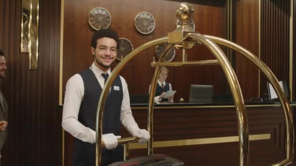ホテルのロビーに立って仕事でカメラを見ている若い笑顔のポーターの中央ショット — ストック動画