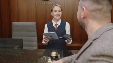 Omuz manzarası üzerinden, otel resepsiyonuna giriş yaparken erkek müşterilerin pasaport bilgilerini dijital tablet üzerinden giriş yapan neşeli genç otel yöneticisi