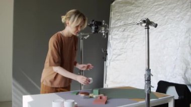 Stüdyoda çalışan genç beyaz kadın fotoğrafçının yatay rafta kamerayla flatlay çekerken ve masa üzerindeki geometrik şekilleri ayarlarken orta boy görüntüsü.