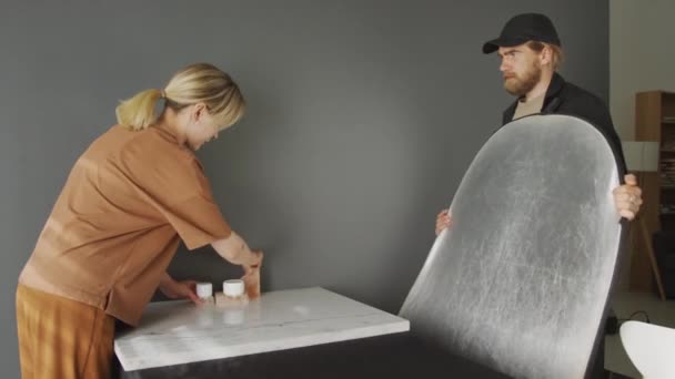 白种人的年轻女子在桌上摆弄一壶手工制作的面霜和装饰性岩石拍照 而男人则站在旁边拿着大的反射镜屏风 — 图库视频影像