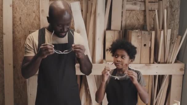 在木制背景的木工工作室工作前 正中镜头的非裔美国人父亲和儿子戴着防护眼镜 — 图库视频影像