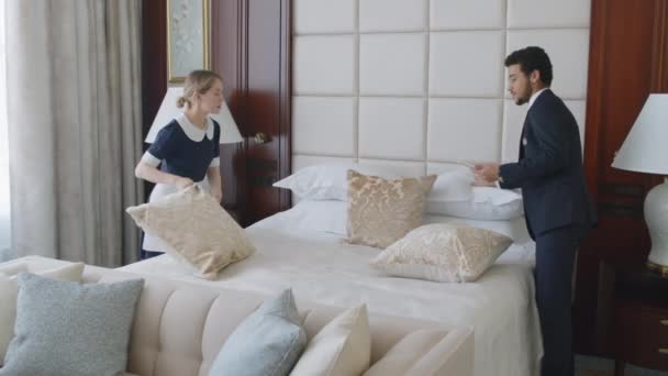 ホテルのマネージャーの中央ショットは 部屋で彼の手でタブレットを保持するベッドを構成するメイドを助けます — ストック動画