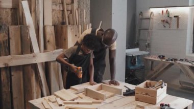 Orta uzunluktaki Afrikalı Amerikalı çocuğun matkapla delik açtığı, babasının marangozluk atölyesinde birlikte çalışırken ona yardım ettiği fotoğraf.