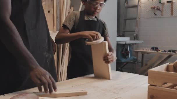 アフリカ系アメリカ人の父親が 彼の息子がワークショップで一緒に作ったときに木製の板を磨くのを手伝って撮影した — ストック動画