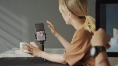 Genç bir kadının, reklam amaçlı yüz kremi örneklerini çekerken, akıllı telefonu tripodda kullanırken ve tanınmayan bir erkek arkadaş tavsiyesi isterken orta seçmeli odak fotoğrafı.