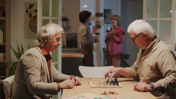 ホームパーティーの背景でワインを飲んでいる間 チェスを演じる濃縮された高齢男性の中型ショット — ストック動画