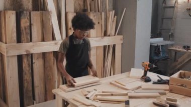 Afro-Amerikalı çocuk marangozluk atölyesinde çalışırken masaya tahta koyuyor ve tek başına deliyor.