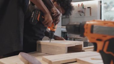 Afro-Amerikalı yetişkin uzman, marangozluk atölyesinde kuş evi yaparken, çocuğun ahşap tahtalara delikler açmasına yardım ediyor.